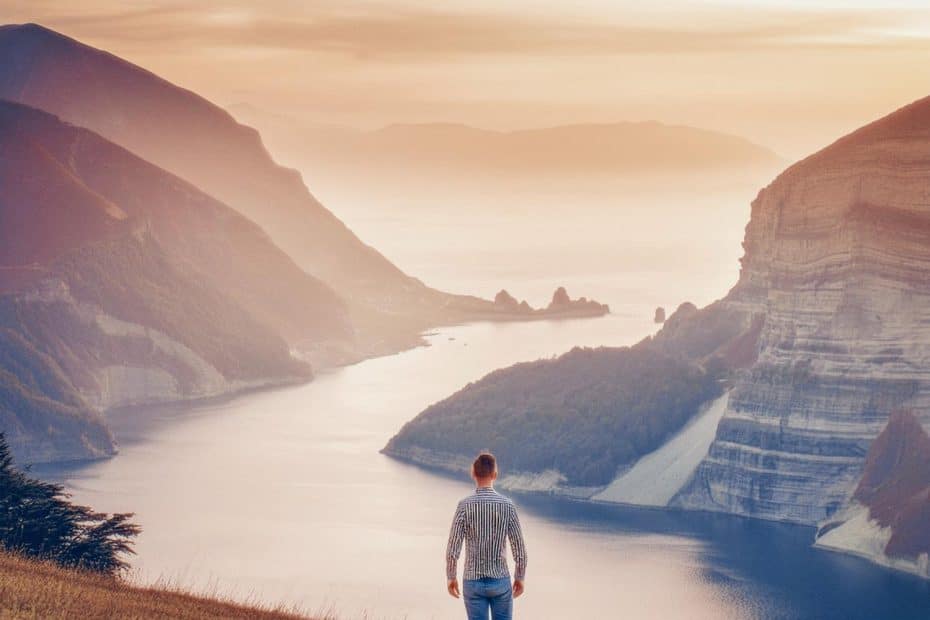 A hiker stands on a grassy hill, gazing at a tranquil lake nestled among steep cliffs and rugged mountains. The sky teems with soft oranges and pinks of sunset, casting a serene glow over the wilderness. This breathtaking vista is an adventurer's paradise, offering an awe-inspiring reward for those who brave the trails to discover nature's hidden gems.