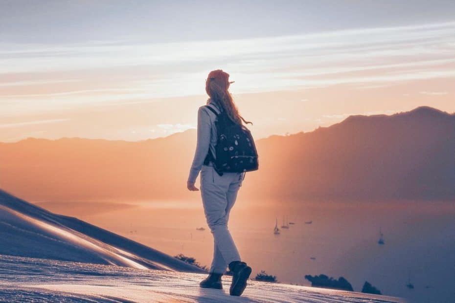 A hiker, adorned in a red cap and white attire, equipped with a backpack, stands on a snow-blanketed slope. They gaze out over a tranquil body of water dotted with boats, encased by majestic mountains bathed in the warm glow of sunset. As the sky transitions from vibrant orange to calming blue tones, they contemplate essential hiking etiquette practices that enable all adventurers to enjoy the great outdoors harmoniously.