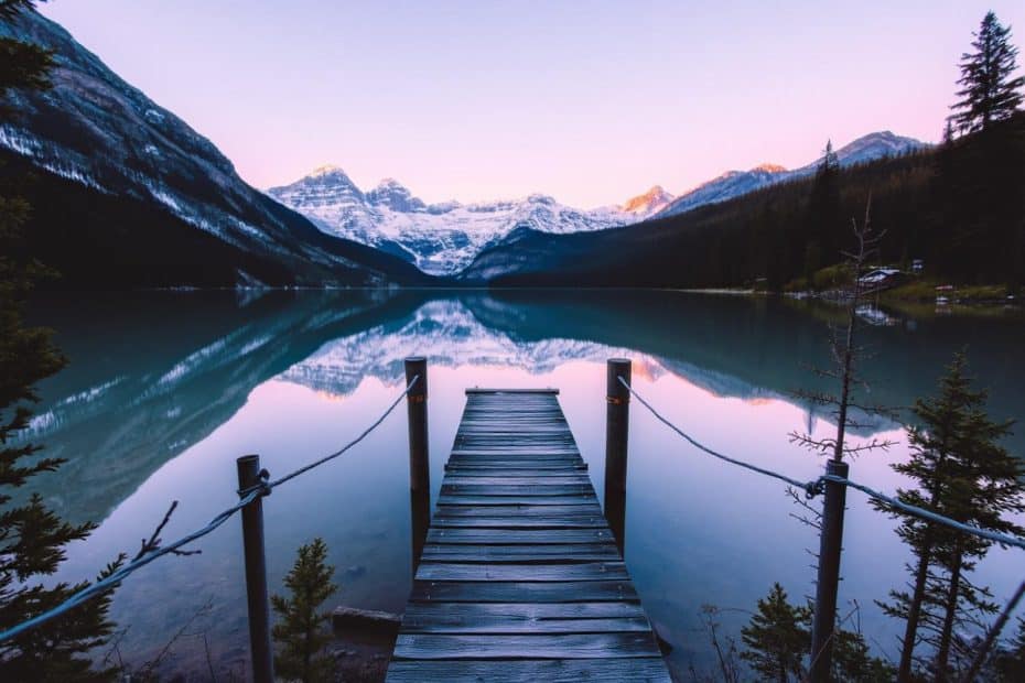 A weathered wooden pier stretches out over a serene, glass-like lake, perfectly mirroring towering snow-capped mountains and the soft glow of a dusky sky. Majestic evergreen trees stand sentry along the shore, completing this tranquil wilderness vista. Imagine setting up camp nearby, with the promise of peaceful hikes and breathtaking sunsets in this idyllic outdoor haven.
