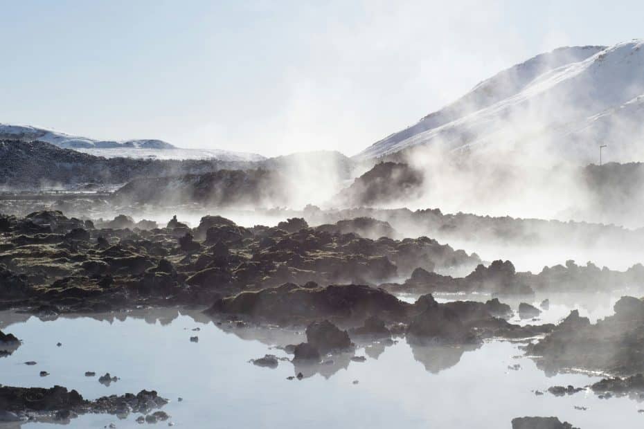 Billowing steam drifts from rocky ground by a mirrored pool, framed by snowy hills—a perfect hot tent camping scene in winter.
