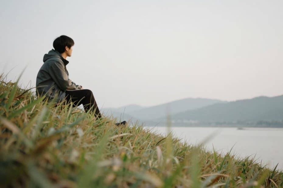 A lone hiker rests on a lush grassy slope beside a gently flowing river, gazing thoughtfully at rolling distant hills beneath a soft, pale sky—capturing a peaceful moment of reflection during an outdoor adventure.